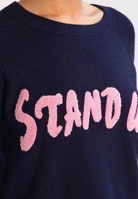 Marinblå sweatshirt med en texturerad rosa slogan "STAND UP". Har en ribbad halsringning och raglanärmar för en avslappnad passform.