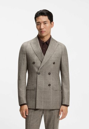 Man draagt een lichtbruine dubbelgeknoopte pak en een donkerbruine shirt staande tegen een effen witte achtergrond.