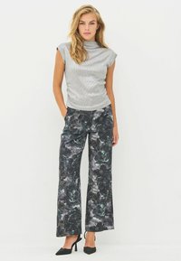 Žena s plavom kosom nosi srebrni kratki top s kratkim rukavima, crne široke hlače s cvjetnim uzorkom i crne cipele s visokom petom, stojeći pred bijelom pozadinom.