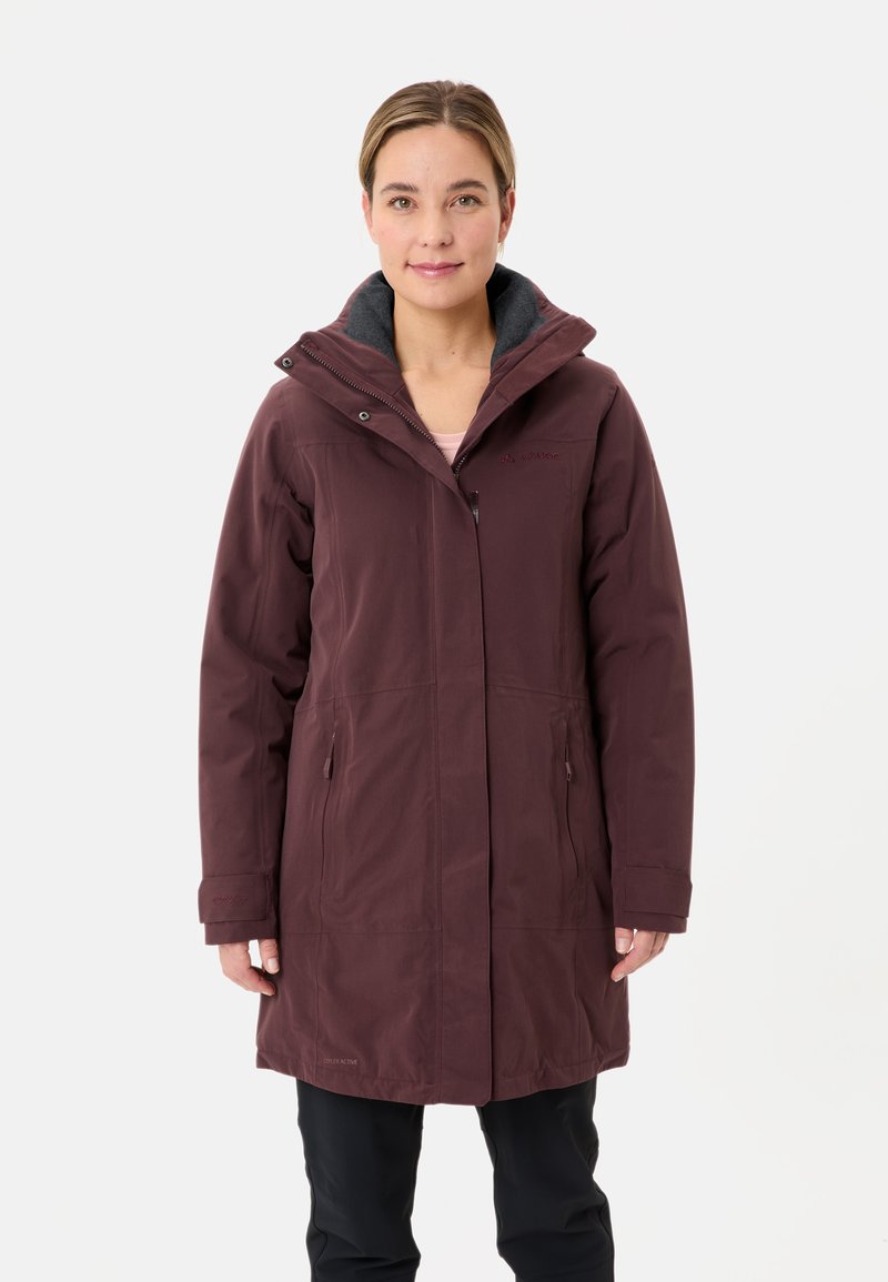 Vrouw in een lange, bordeauxrode, met een capuchon winterjas met ritssluiting zakken en zwarte broek, staande tegen een effen witte achtergrond.