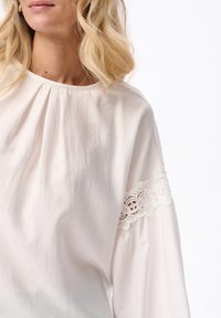 Blondynka w luźnej białej bluzce z koronkowymi detalami na rękawach i delikatnymi fałdami przy dekolcie.