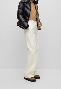 Černá péřová bunda s prošívaným designem, béžový pletený top, krémové kalhoty s širokými nohavicemi a hnědé šněrovací boty s černou podrážkou.