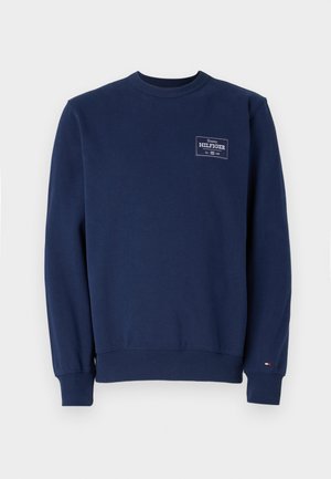 Tummansininen puuvillainen collegepaita, jossa on pyöreä kaula-aukko ja ribbineulos hihansuissa ja helmoissa. Vasemmassa rinnassa on valkoinen logotarra. Sujuva pinta.