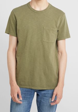 Homme portant un t-shirt vert olive à manches courtes, col rond avec poche poitrine, et un jean bleu déchiré sur fond uni.