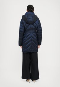 Vrouw die met haar rug naar ons toe staat, gekleed in een marineblauwe gewatteerde jas met capuchon, wijde zwarte broek en beige hoge hakken, tegen een effen achtergrond.