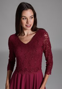 Robe bordeaux avec un corsage en dentelle ornée de motifs floraux et de sequins, décolleté en V, et manches en dentelle trois-quarts ; jupe fluide.