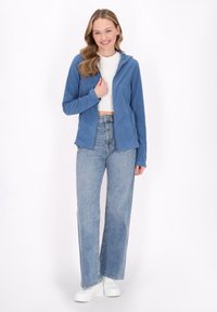 Felpa blu con zip realizzata in tessuto morbido, con cappuccio e tasche frontali, abbinata a jeans chiari a vita alta e sneakers bianche.