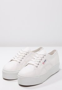 Baskets en toile blanche à surface texturée, lacets plats et semelle compensée épaisse. Présentent un logo rouge et bleu sur le côté.