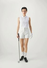 Λευκό αμάνικο πόλο μπλουζάκι, κομψές λευκές σορτς, μαύρα παπούτσια γκολφ, λευκό γάντι γκολφ και κάλτσες, κρατώντας ένα μπαστούνι γκολφ. Ουδέτερο φόντο.