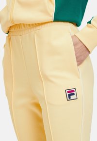 Gele trainingsbroek van zacht materiaal met zijzakken en witte bies. Heeft een kleurrijke FILA-logo patch op de bovenbeen.