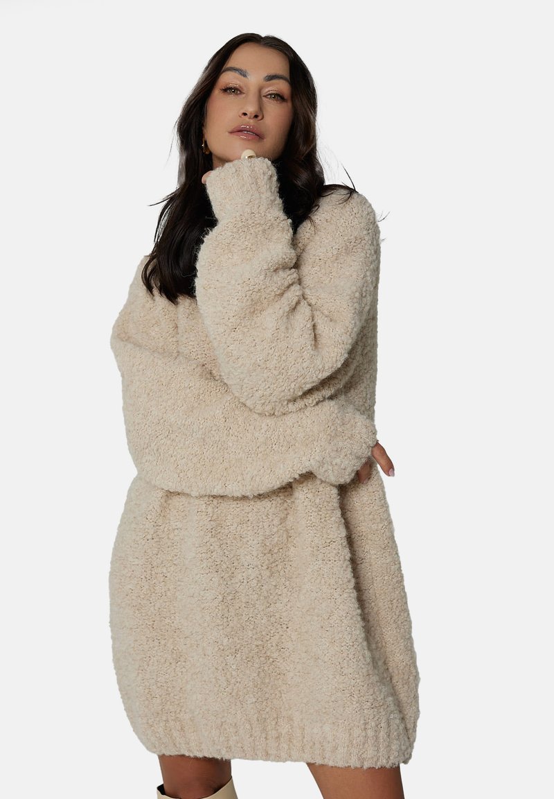 Puszysta beżowa oversize'owa sukienka sweterowa z wysokim kołnierzem, długimi rękawami i prążkowanym dołem, charakteryzująca się miękką teksturą i luźnym krojem.