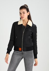 Giacca bomber nera con colletto in pelliccia beige, chiusura con zip frontale, polsini a coste e un'etichetta arancione sull'orlo. Presenta tasche laterali e nelle maniche.