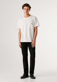 Camiseta blanca de algodón con un bolsillo en el pecho que lleva texto, combinada con pantalones negros y zapatos negros. Diseño sencillo y clásico.