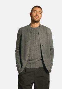 Šedý vlněný cardigan s kulatým výstřihem, zapínáním na knoflíky a žebrovanými manžetami, vrstvený přes texturovaný šedý svetr a doplněný o tmavé kalhoty.