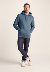 Blue hoodie with kangaroo pocket, drawstring hood, and logo detail. Worn with dark denim jeans and white sneakers with black stripes.