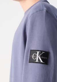 Zbliżenie osoby noszącej jasnofioletową bluzę z naszywką z czarnym logo Calvin Klein Jeans na górnej części ramienia.