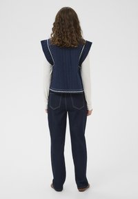 Gilet imbottito blu navy con spalle arricciate, dettaglio di rifinitura bianca. Abbinato a pantaloni di jeans blu scuro e calzature marroni.