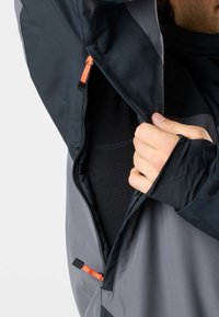 Person partially unzipping a grey and black jacket, revealing a black inner layer, with orange zipper pulls and thumb hole in the sleeve.