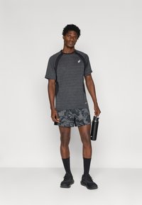 Αθλητική εμφάνιση με γκρι μπλουζάκι κοντομάνικο με μαύρες λεπτομέρειες και μαύρη σορτς με σχέδιο. Το μοντέλο κρατάει ένα μαύρο μπουκάλι νερού.