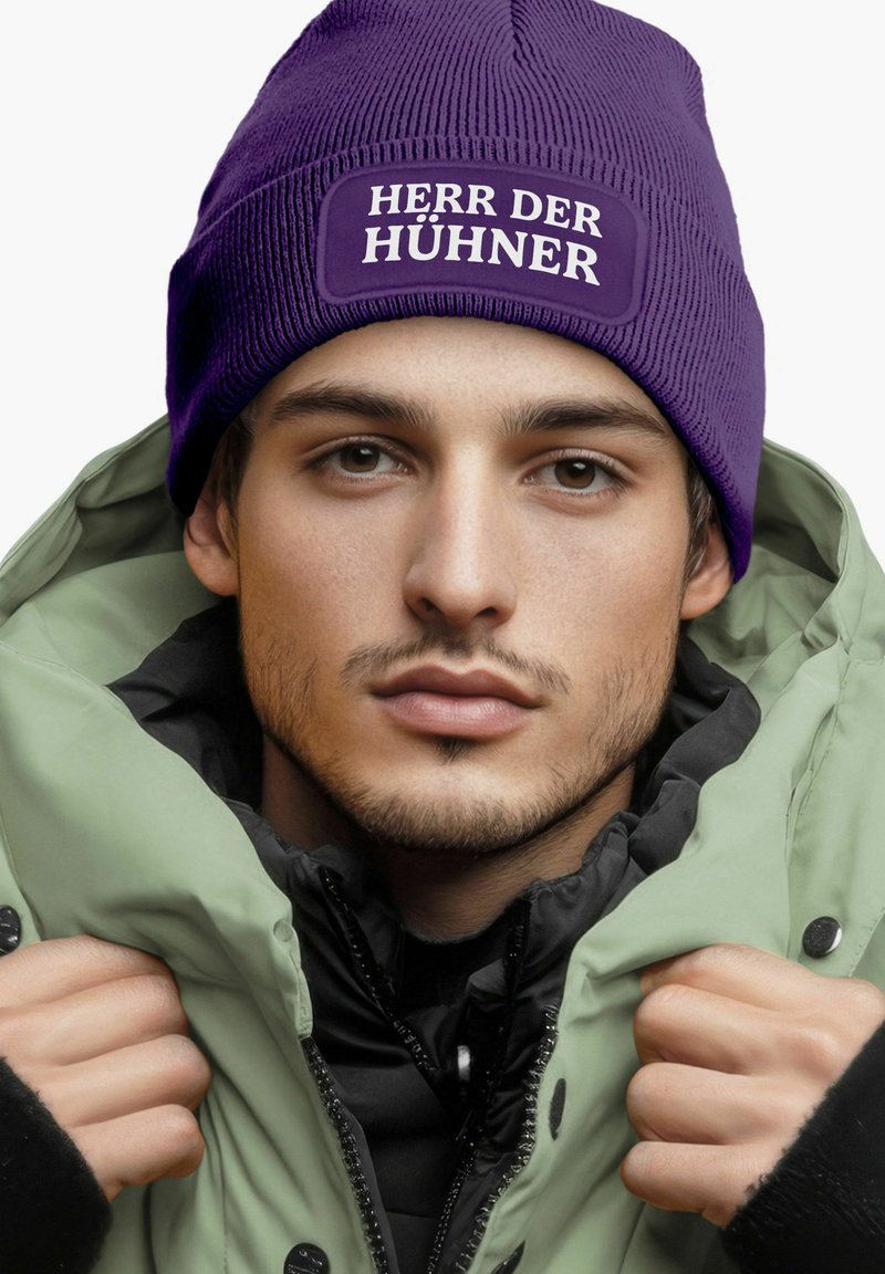 Purple knitted beanie with white text "HERR DER HÜHNER" above a green and black puffer jacket. Soft texture, snug fit, and layered design.