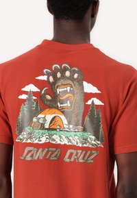 Orange cotton T-shirt featuring a graphic of a large bear's paw, a tent, and trees, with "SANTA CRUZ" printed at the bottom in a wood texture.