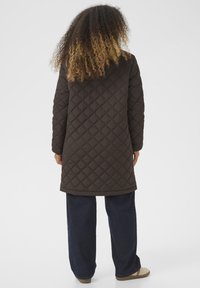 Manteau long matelassé marron avec capuche, arborant un motif en losanges. Associé à un pantalon large bleu foncé et des chaussures beiges.