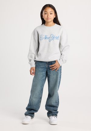 Fille aux longs cheveux noirs portant un sweat-shirt gris "New York", un jean bleu ample et des baskets blanches, se tenant contre un fond uni.