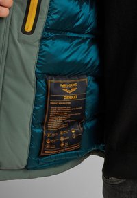 Giacca corta con cappuccio in ripstop verde con interno blu, dotata di polsini regolabili e un'etichetta del prodotto che mostra le specifiche in testo nero e oro.