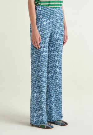 Donna che indossa pantaloni a zampa larghi a vita alta di colore blu con motivo geometrico bianco e verde, abbinati a ballerine con punta a mosaico.