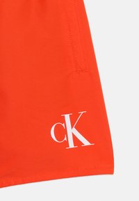 Λαμπερό πορτοκαλί ύφασμα με λευκό λογότυπο «cK» κοντά στην κάτω δεξιά γωνία και κάθετη ραφή ραμμένη πάνω από το λογότυπο.