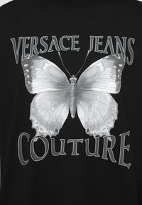 Μαύρο μπλουζάκι με μεγάλη ασημένια γραφική εικόνα πεταλούδας, με το κείμενο "VERSACE JEANS COUTURE" σε αντίθετη ασημένια γραμματοσειρά από πάνω και από κάτω.