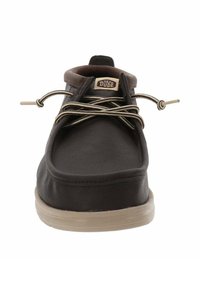 Scarpa slip-on in pelle marrone con suola in gomma beige, punta tondeggiante e lacci decorativi color cammello. Presenta un logo "Hey Dude" al centro superiore.