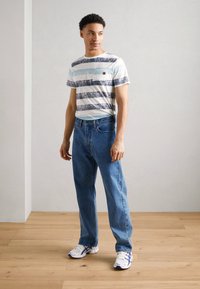 Camiseta de algodón a rayas en blanco, azul y marino; jeans de denim azul claro; zapatillas deportivas blancas con detalles azules; entorno interior con suelo de madera clara.