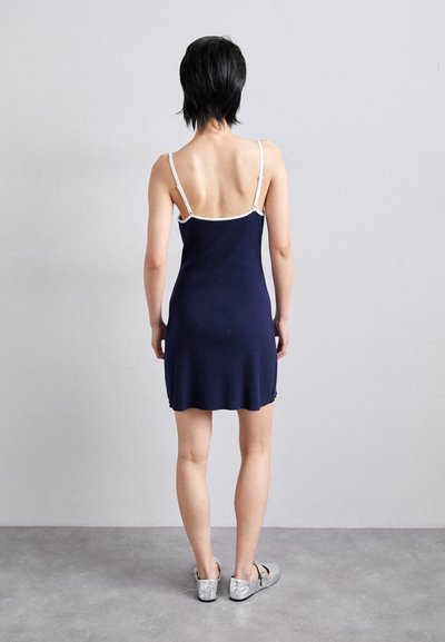 Vestido de punto azul marino con silueta ajustada, tirantes finos blancos y un dobladillo que cae a medio muslo. Se lleva con zapatos metálicos plateados.