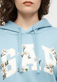 Κοντινό πλάνο ενός ατόμου που φοράει ανοιχτό μπλε φούτερ με κουκούλα, με λευκά γράμματα "GAP" διακοσμημένα με σχέδια από λουλούδια μαργαρίτας.