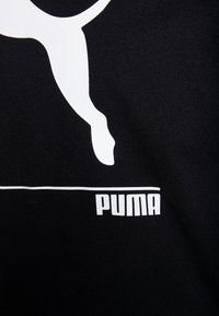 Logo Puma blanc d'un chat sautant au-dessus du mot Puma sur un fond en tissu noir.