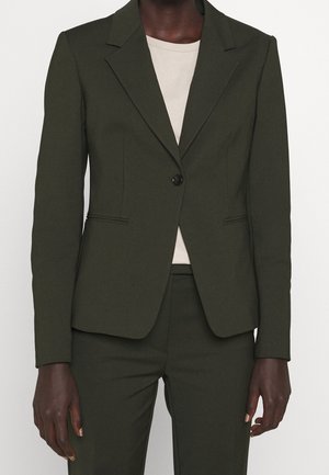 Donkergroene op maat gemaakte blazer met één knoopsluiting gedragen over een lichtbeige top met ronde hals, gecombineerd met bijpassende donkergroene broek.