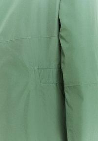 Grüne leichte Jacke mit glatter Textur, elastischen Details an der Taille und einem klaren, minimalistischen Design ohne sichtbare Verschlüsse.