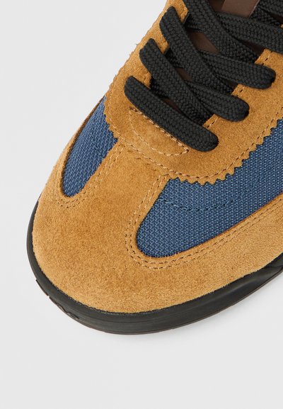 Brūnas zamšādas sporta kurpes ar zilām acs detaļām un melnām kurpju šņorēm. Aprīkotas ar apaļu purngalu, fakturētiem materiāliem un gumijas zolīti pret slīdēšanu.