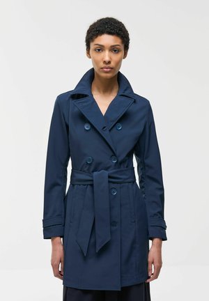 Kvinde stående iført en dobbeltradet navy trenchcoat med bindebælte og bred krave mod en ensartet lys baggrund.