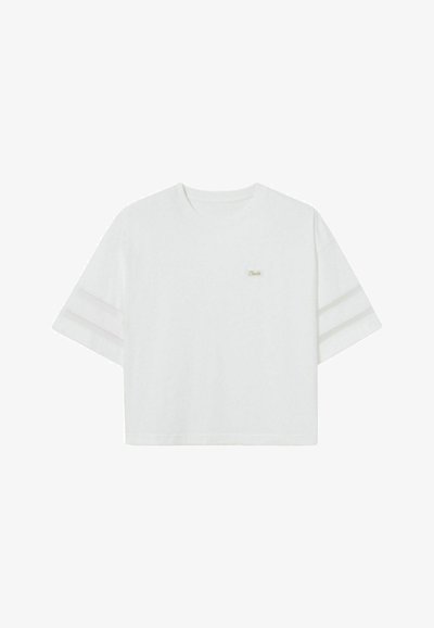 T-shirt blanc court fabriqué en tissu doux, avec des manches courtes et trois rayures horizontales sur chaque manche. Petit logo sur la poitrine.