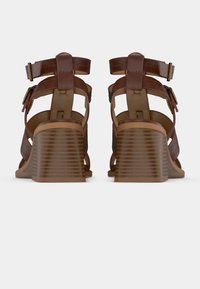 Ζευγάρι καφέ δερμάτινα σανδάλια με λουράκια, με χοντρά τακούνια και κλείσιμο με αγκράφα στον αστράγαλο, εμφανιζόμενα από πίσω σε λευκό φόντο.