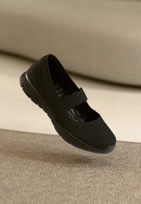 Black Mary Jane-style shoe with a fabric upper, elastic strap, and textured sole, featuring a breathable design and minimalist shape.