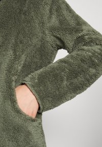 Olivgrön plyschfleecejacka med en mjuk, fluffig textur, vida ärmar och en sidoficka med en rundad öppning.