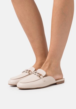 Pieds portant des mocassins beiges sans talon avec un détail en chaîne dorée et beige sur fond blanc.