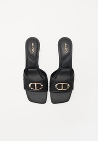 Sandales en plastique noires avec une sangle texturée, dotées d'une boucle dorée, semelle plate et semelle intérieure rembourrée pour le confort. Logo de la marque visible.