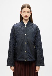 Tengerészkék steppelt dzseki álló gallérral, arany patentgombokkal és lelógó vállrésszel. Simább, könnyű anyagból készült.