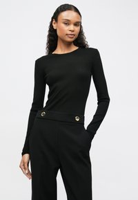 Jovem mulher com cabelo alongado e encaracolado, vestindo uma blusa preta de manga longa com textura canelada e calças altas pretas com botões dourados, com as mãos nos bolsos.