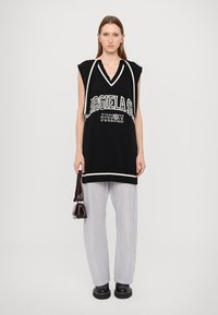 Top de malha preto sem mangas com um decote em V e acabamentos brancos, apresentando texto e números. Combinado com calças claras às riscas e botas pretas.