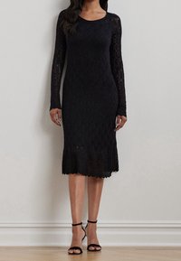 Czarna koronkowa sukienka z długimi rękawami, teksturowanym wzorem, dopasowanym krojem, do kolan oraz falbaniastymi krawędziami, zestawiona z czarnymi sandałkami na paski.